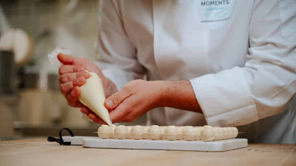 Man sieht die Hände eines Konditors mit einem Spritzbeutel mit weißer Schokolade, der weiße Schokoladenrohlinge befüllt