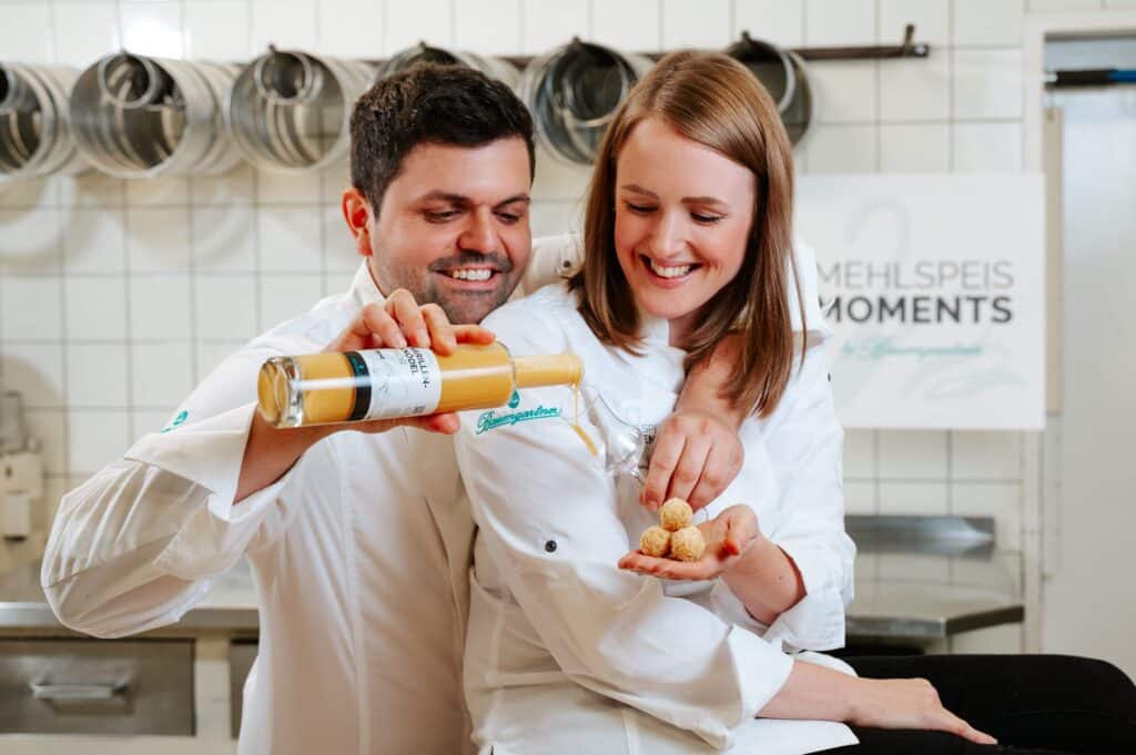 Julia und Mathias Baumgartner in der Backstube - Julia hält Pralinen und ein Schnapsglas und Mathias schenkt in das Glas ein. Beide lachen herzlich
