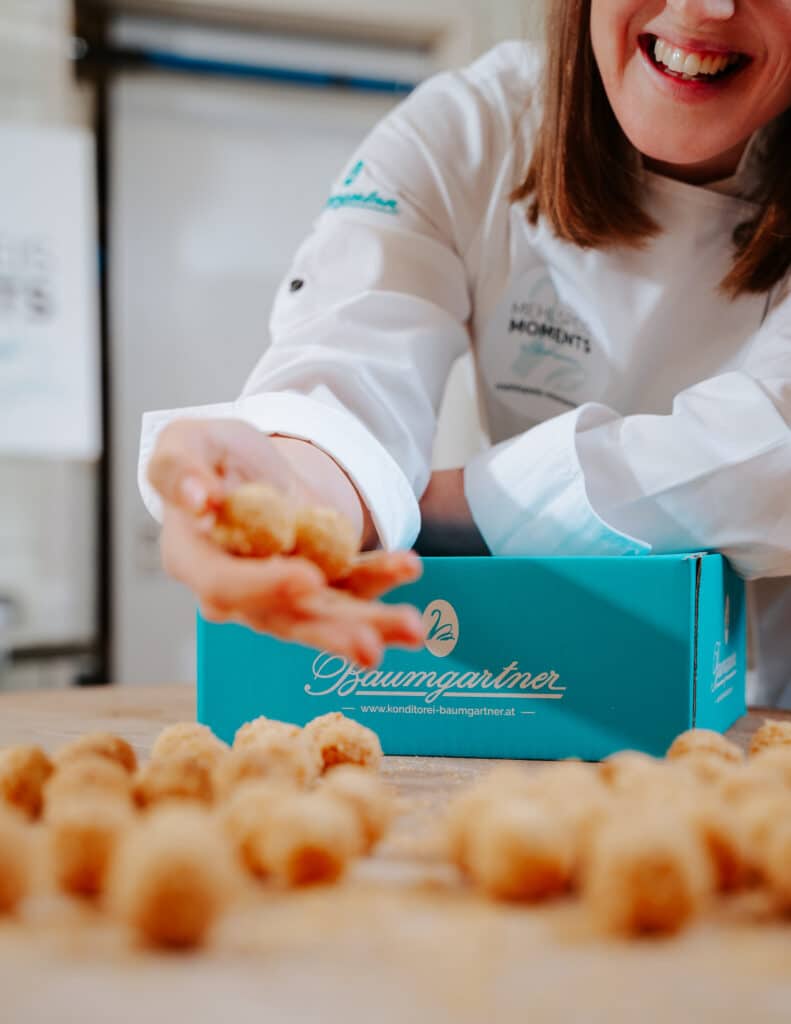 Marillenknödel Pralinen im Vordergrund, dahinter Julia Baumgartner mit einer Versandbox in Türkis von Baumgartner